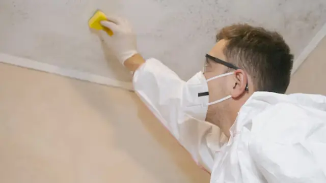 Person in a mask and gloves wiping a mold-spotted ceiling corner with a sponge